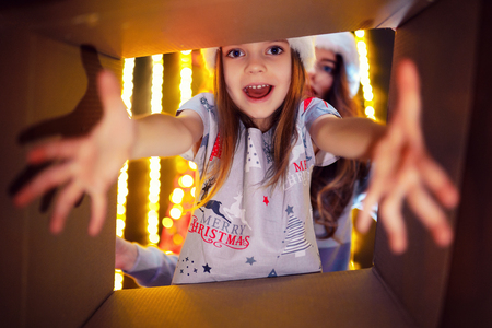 Cheerful mom and her cute daughter girl opening a Christmas present. Parent and little child having fun near Christmas tree indoors. View from inside of the box.の写真素材