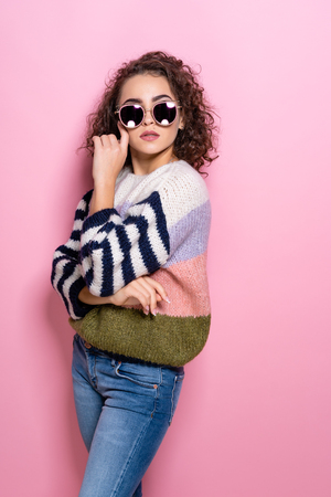 Beautiful girl with curly hair posing in fashionable sweater, jeans and sunglasses. Young girl standing on pink pastel background.の写真素材