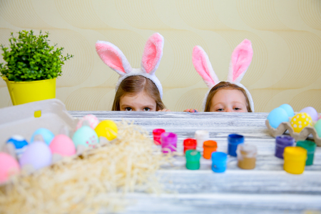 Curious small girls in bunny ears peeking out from behind kitchen table with tasty easter eggs and looking at camera with surprise. Focus on childrenの写真素材