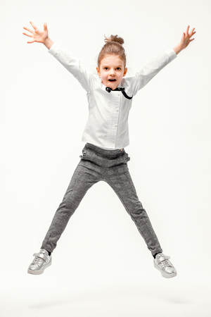 Beautiful little girl dressed like a School girl - in white shirt and gray pants, rounded glasses standing near white wall and posing, jumping like model.の写真素材
