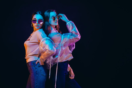 Side view of provocative teen girls in stylish jackets and sunglasses standing on black background in neon illuminationの写真素材