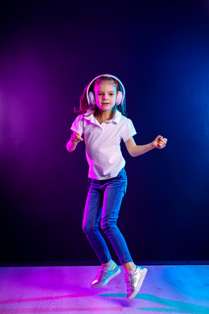 Girl of 7 years old listening to music in headphones and jump on dark colorful background . Dancing girl. Happy small girl dancing to music. Cute child enjoying happy dance music.の写真素材