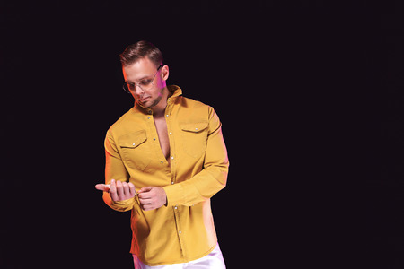 Positive young male looking away and adjusting trendy yellow shirt while standing under colorful...