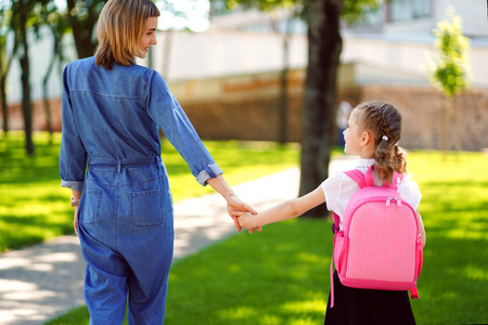 Parent and pupil of primary school go hand in hand. Woman and girl with pink backpack behind the back. Beginning of lessons. First day of fallの写真素材