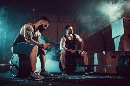 Two muscular bearded tattoed athletes relax after training, lifting heavy weight. smoke at gym. Scream. Working hard. Exercise for the muscles of the backの写真素材