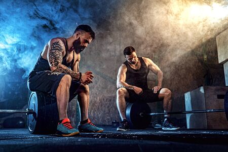 Two muscular bearded tattoed athletes relax after training, lifting heavy weight. smoke at gym. Scream. Working hard. Exercise for the muscles of the backの写真素材