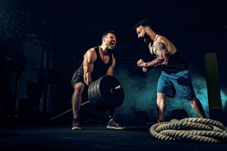 Two muscular bearded tattoed athletes training, one lift heavy weight bar when other is motivating. Scream. Working hard. Exercise for the muscles of the backの写真素材