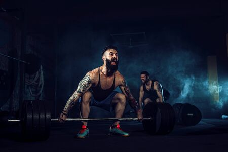 Two muscular bearded tattoed athletes training, one lift heavy weight bar when other is motivating....