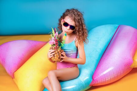 Pretty smiling girl wearing pink and blue swimwear and sunglasses holding pineapple cocktail with colorful straws and showing thumb up on rainbow inflatable mattress backgroundの写真素材