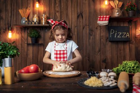 Smiling girl wearing white T-shirt with checkered apron and headband kneading bread dough on table filled with ingredients for pizza in stylish wooden kitchen.の写真素材