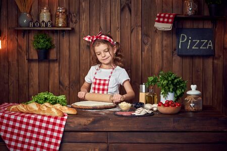 Smiling girl wearing white T-shirt with checkered apron and headband preparing dough with roller pin on table filled with ingredients for pizza, looking at camera in stylish wooden kitchen.の写真素材