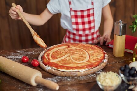 Girl wearing checkered apron and headband cooking pizza by spreading sauce with spoon in heart form on table filled with ingredients for pizza, in stylish wooden kitchen.の写真素材