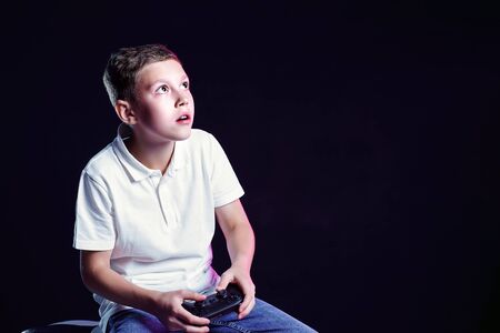 Cheerful boy in white t-shirt with game pad in hands playing and looking away from camera on black...