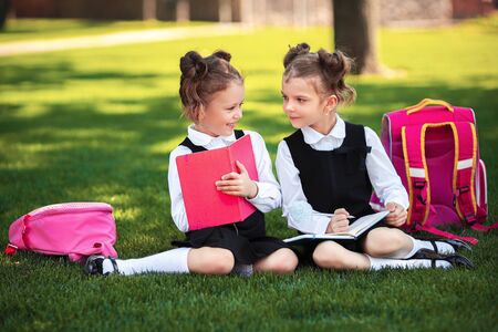 Two little school girls with pink backpack sitting on grass after lessons and read book or study...