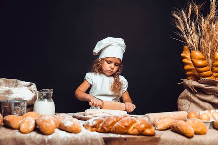 Charming little girl with curly hair in white apron and hat standing at table kneading bread dough and looking at camera. KId in good mood, having fun.の写真素材