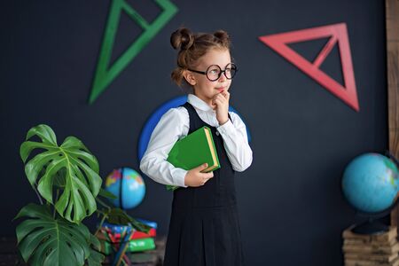Clever little girl in school uniform and glasses holding green textbook before studiesの写真素材