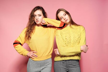 Two beautiful sexy smiling gorgeous girls looking at camera. Hot women standing in stylish yellow sweaters, on pink background. Studio shot. Isolateの写真素材