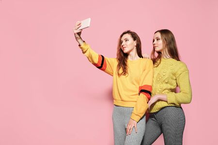 Two cheerful young women with listening to music and talking selfie over pink background