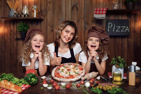 Cheerful woman with cute girls in aprons and hats smiling and looking at camera show thumb up during pizza preparation in kitchen at homeの写真素材