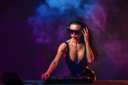 Young sexy woman dj playing music. Headphones and dj mixer on table. Colorful Smoke on background