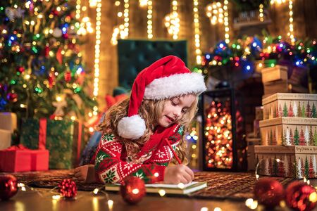Little charming girl in Santa hat is lying on the floor and writes a letter, draws with a pencil. lights of garlands. warm comfortable roomの写真素材
