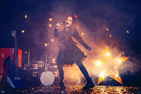 Beautiful girl with curly hair wearing leather jacket, boots sing into a wireless microphone for karaoke like rock star in recording studio or stage. Smoke on background.の写真素材