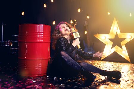 Beautiful girl with curly hair singing into a wireless microphone for karaoke leaning on a red tank in recording studio.の写真素材