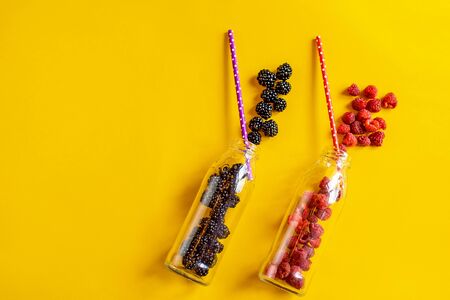 Blackberry and raspberry fruit in glass bottles with straws on yellow background. Fresh organic...