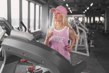 Side view of sporty blonde woman in pink cap exercising on treadmill in gym.