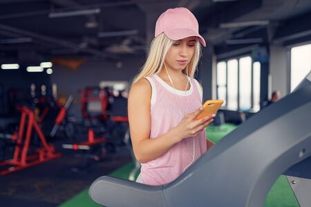 Side view of sporty blonde woman in pink cap prepearing for exercising on treadmill by choosing...