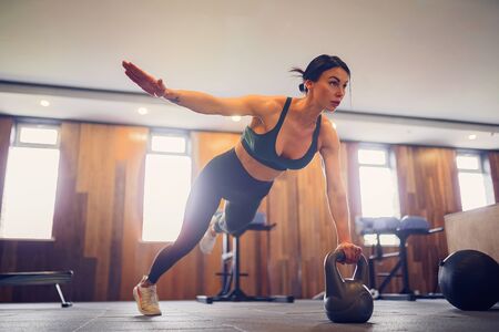 young motivated girl doing plank exercise using kettlebells with one hand at gym, full length photo, copy space.の写真素材