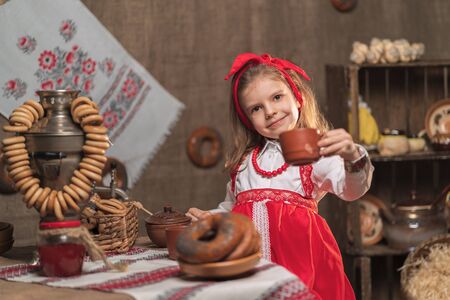 Little girl wearing red headband and ornamental shirt drinking tea from samovar celebrating Maslenitsaの写真素材