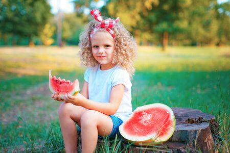 Happy curly child girl sitting on stump and eating watermelon outdoors in spring park against natural sunny blurred background.の写真素材