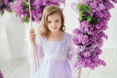 Beautiful little girl of preschool age in a delicate dress sitting on a swing decorated with flowers of lilac.の写真素材