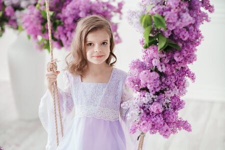Beautiful little girl of preschool age in a delicate dress sitting on a swing decorated with flowers of lilac.の写真素材