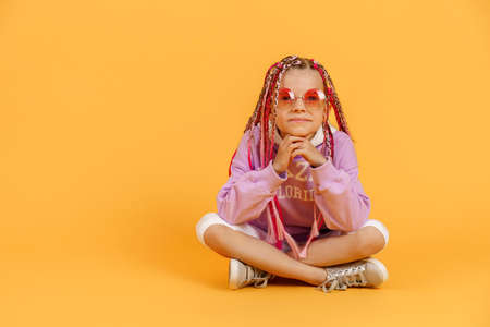 Stylish girl in rounded glasses with pink dreadlocks sitting, looking at camera and posing on a yellow background. Beauty, fashion.の写真素材