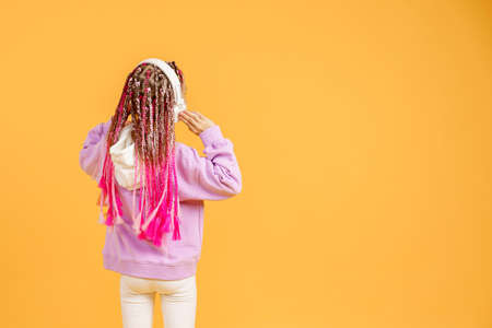 Back view of Adorable child in rounded glasses with pink dreadlocks listen to music playing in earphones on yellow background. Cute small kid listening to music in headset, copy space.の写真素材