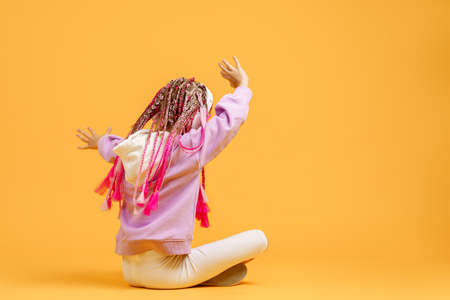 Back view of Adorable child in rounded glasses with pink dreadlocks sitting on floor and listen to...