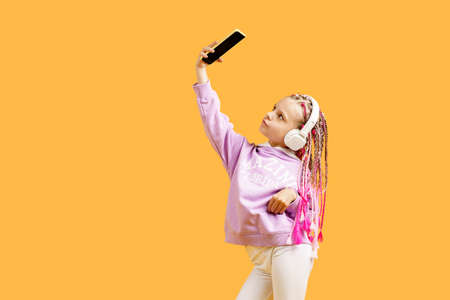 Pretty modern girl with pink dreadlocks in headphones smiling and making selfie on smartphone...
