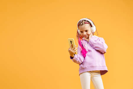 Pretty modern girl with pink dreadlocks in headphones smiling and making selfie on smartphone isolated over yellow.の写真素材
