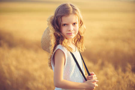 Fashion photo of a little girl in white dress and straw hat at the evening wheat field.