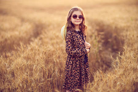 Fashion photo of a little girl in leopard print dress, sunglasses and straw hat at the evening wheat field.の写真素材