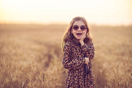 Fashion photo of a little girl in leopard print dress, sunglasses and straw hat at the evening wheat field.の写真素材