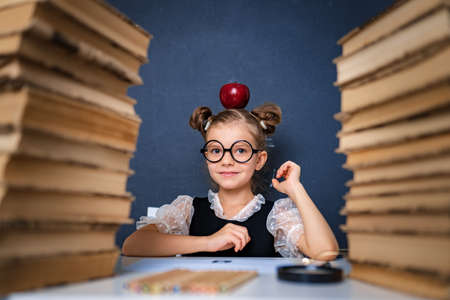 Happy smart girl in rounded glasses sitting between two piles of books with red apple on head and look at camera smiling.の写真素材