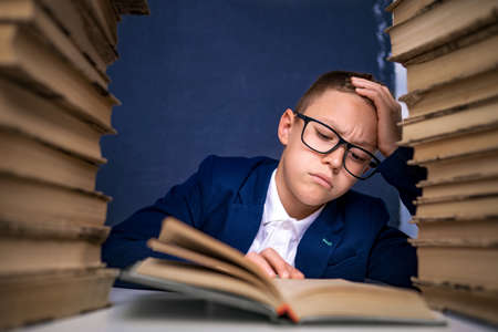 Smart boy in glasses sitting between two piles of books and read book thoughtfully.の写真素材