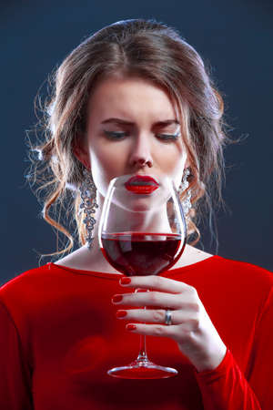 Woman with makeup, hairstyle waering red dress posing with glass of vine over dark background, isolate.の写真素材