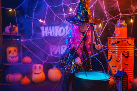 Little beautiful girl in a witch costume celebrates Happy Halloween party in interior with pumpkins.