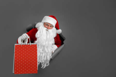 Santa man holds shops packet in hand through a paper hole. Bearded man in santa hat looking through hole on grey paper.の写真素材