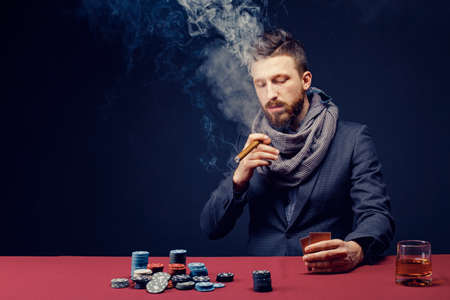 Stylish bearded Man in suit and scarf playing in dark casino, smoking cigar, drink whiskey.の写真素材