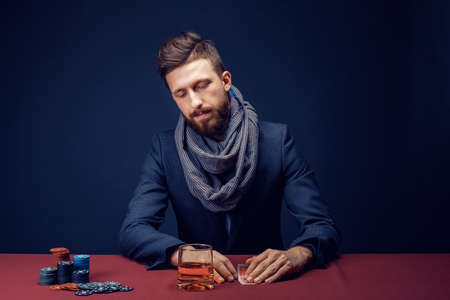 Stylish bearded Man in suit and scarf playing in dark casino, smoking cigar, drink whiskey.